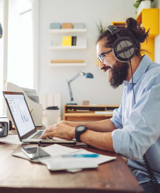 Man wearing headphones and smiling as he types on a laptop in a bright, homey office.