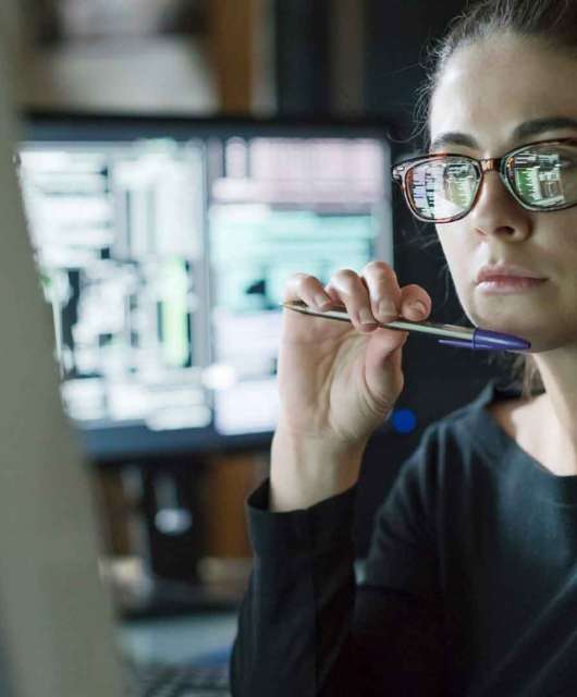 Woman holding pen under her chin looking at a desktop computer.